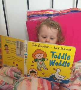 A toddler lays in bed and reads Toddle Waddle, a picture book by Julia Donaldson and Nick Sharratt