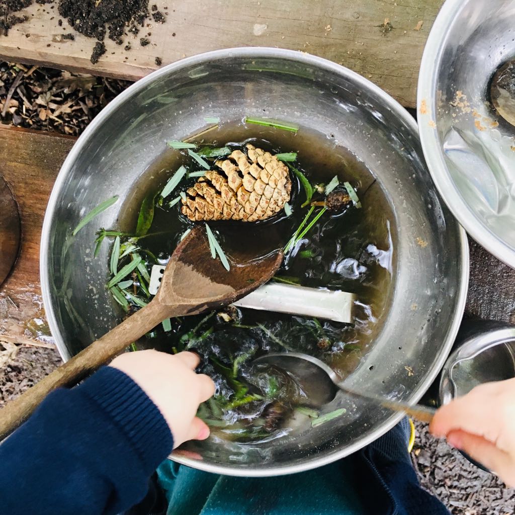 A toddler uses a metal wooden spoon to stir a shallow metal pan filled with muddy water, pine cones and grass clippings during a messy play session at a mud club
