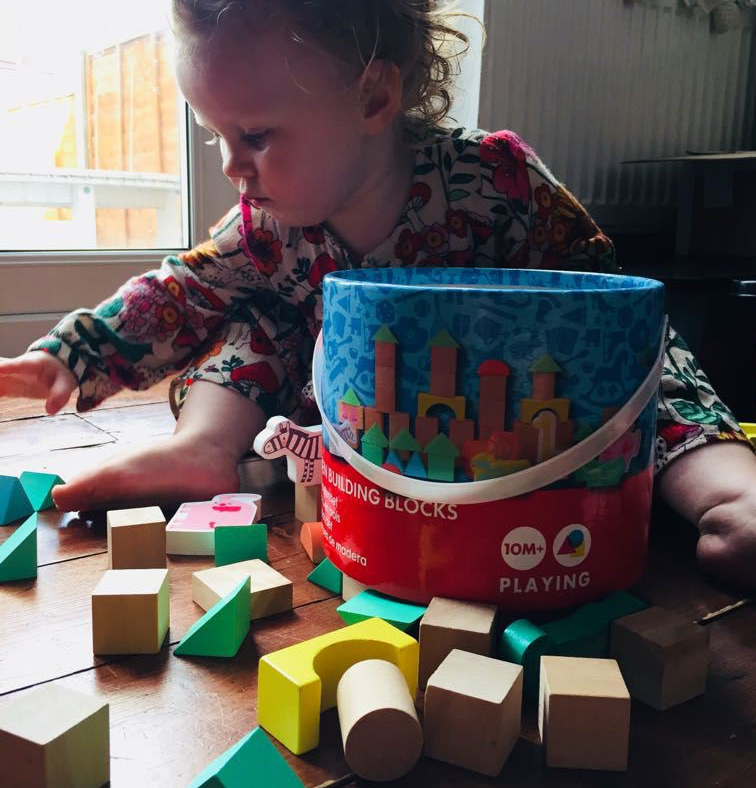 A toddler sits on the floor of a room playing with a bucket of traditional wooden blocks