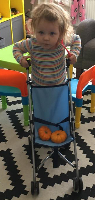 A young child pushing a toy pushchair holding two small pumpkins