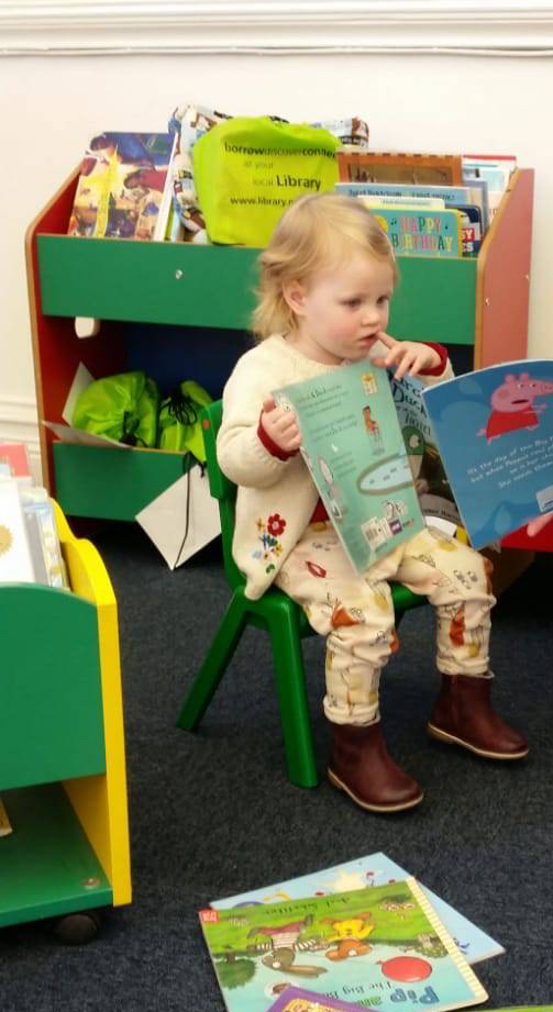A child sitting in a library reading Sarah & Duck and Peppa Pig picture books