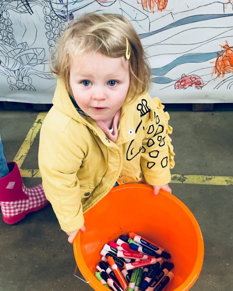 A young child in a yellow coat holding an orange bucket filled with felt tip pens