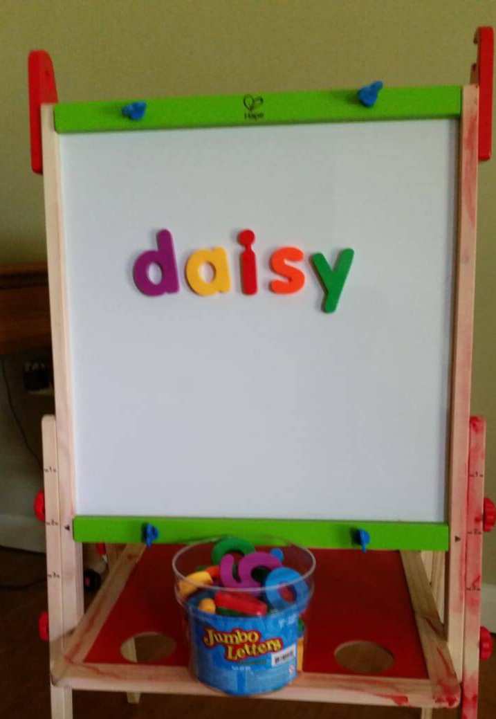 A child's easel with a set of magnetic fridge letters arranged on it to spell the word Daisy