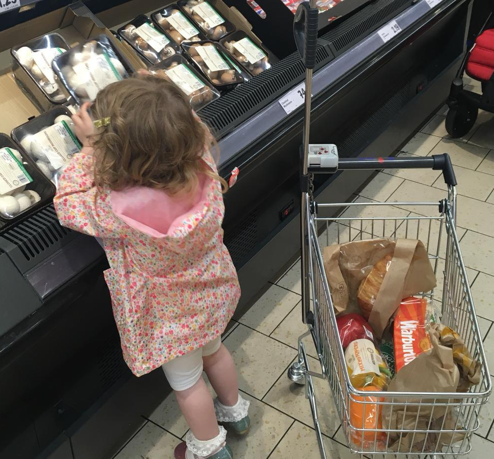 A young child helps her parents select a packet of mushrooms during a shopping trip
