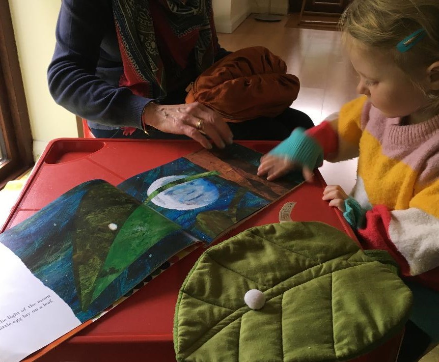 A child reads The Very Hungry Caterpillar by Eric Carle with an adult with the contents of a story sack nearby