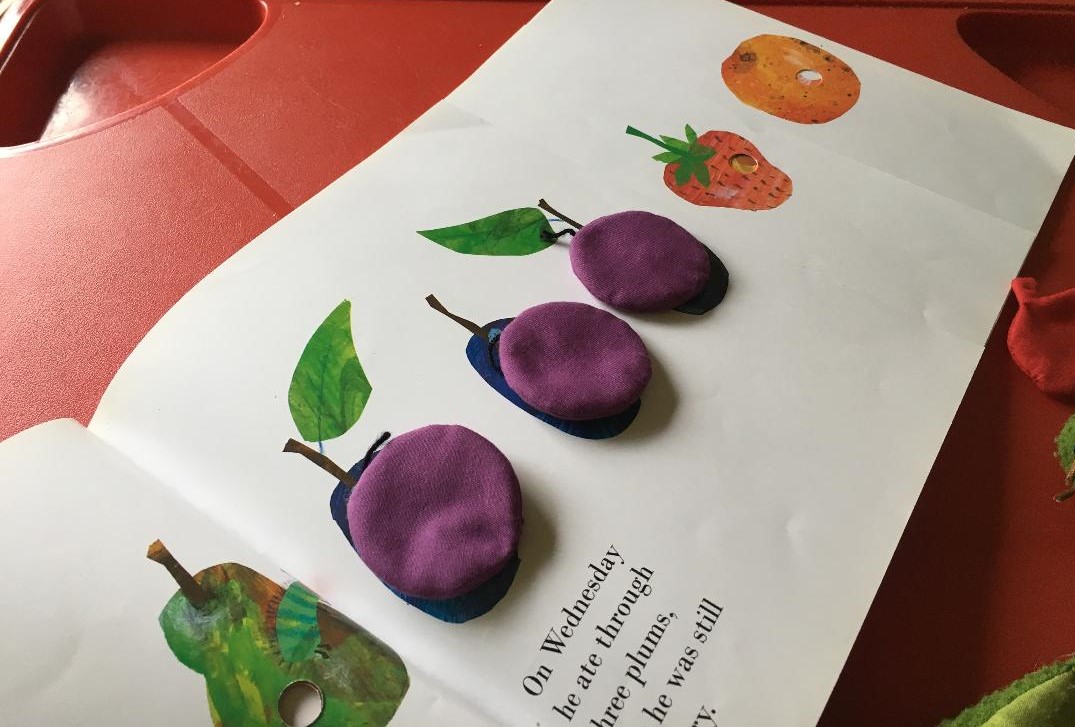 A page from Eric Carle's Very Hungry Caterpillar with soft toy plums from a library story sack laid on top of the corresponding page in the book