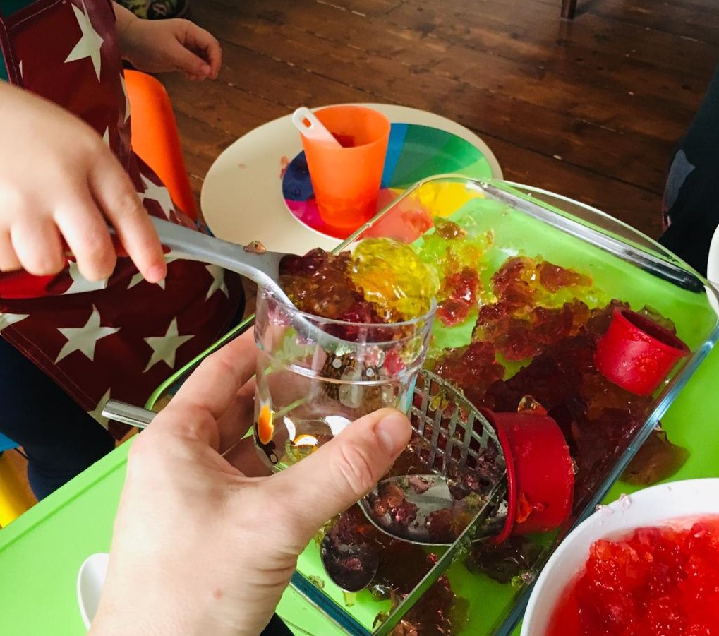 A messy play activity in progress, a child is using an ice cream scoop to serve some jelly (or jello) to an adult who is holding a cup. A potato masher and other cup and cookie cutters are visible.