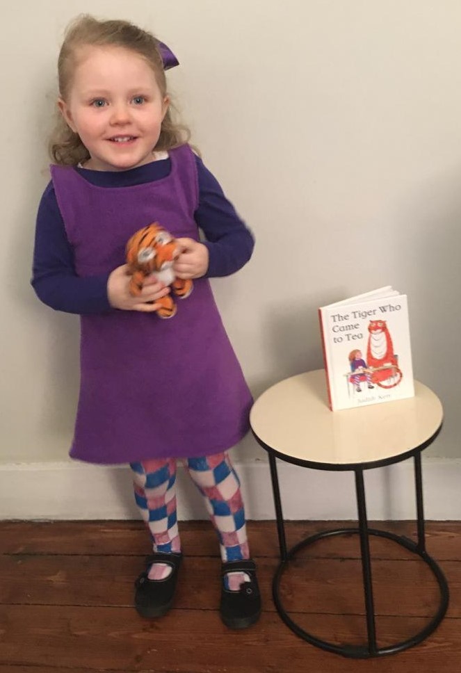 A child dressed as Sophie standing next to a copy of the Tiger Who Came To Tea by Judith Kerr