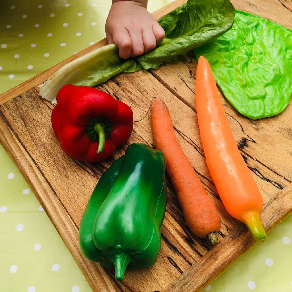 A wooden chopping board show display several vegetables - a pepper, a carrot and a lettuce lead - along with their plastic pretend-play counterparts