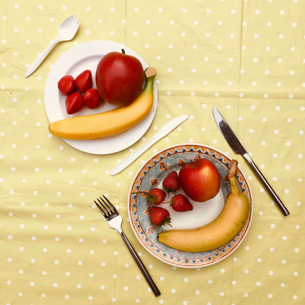 Two plates of food are contrasted, one is a china plate with apple, strawberries and banana, and metal cutlery. The other shows the same arrangement, except the food, crockery and cutlery are all plastic.