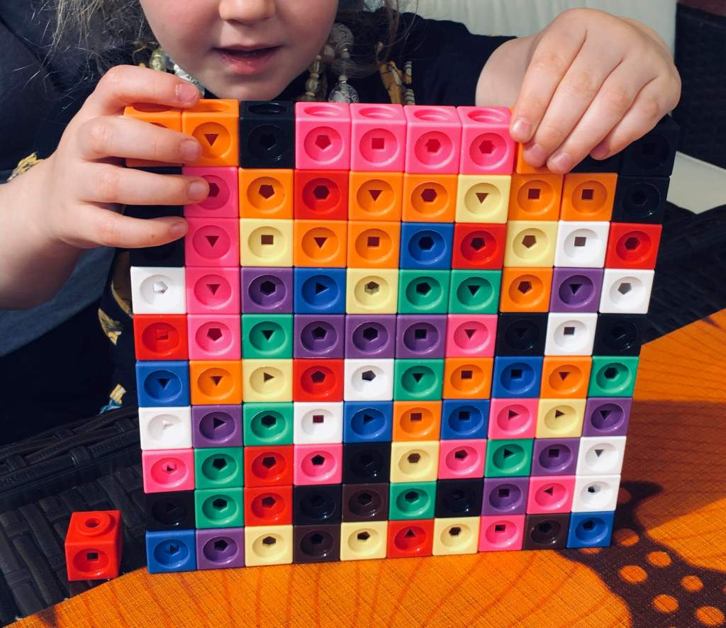 A young girl carefully positions the final cubes in a multicoloured square of Mathlink Cubes, to construct a 10 by 10 square made of 100 individual cubes. A single additional cube is left to one side, for scale.