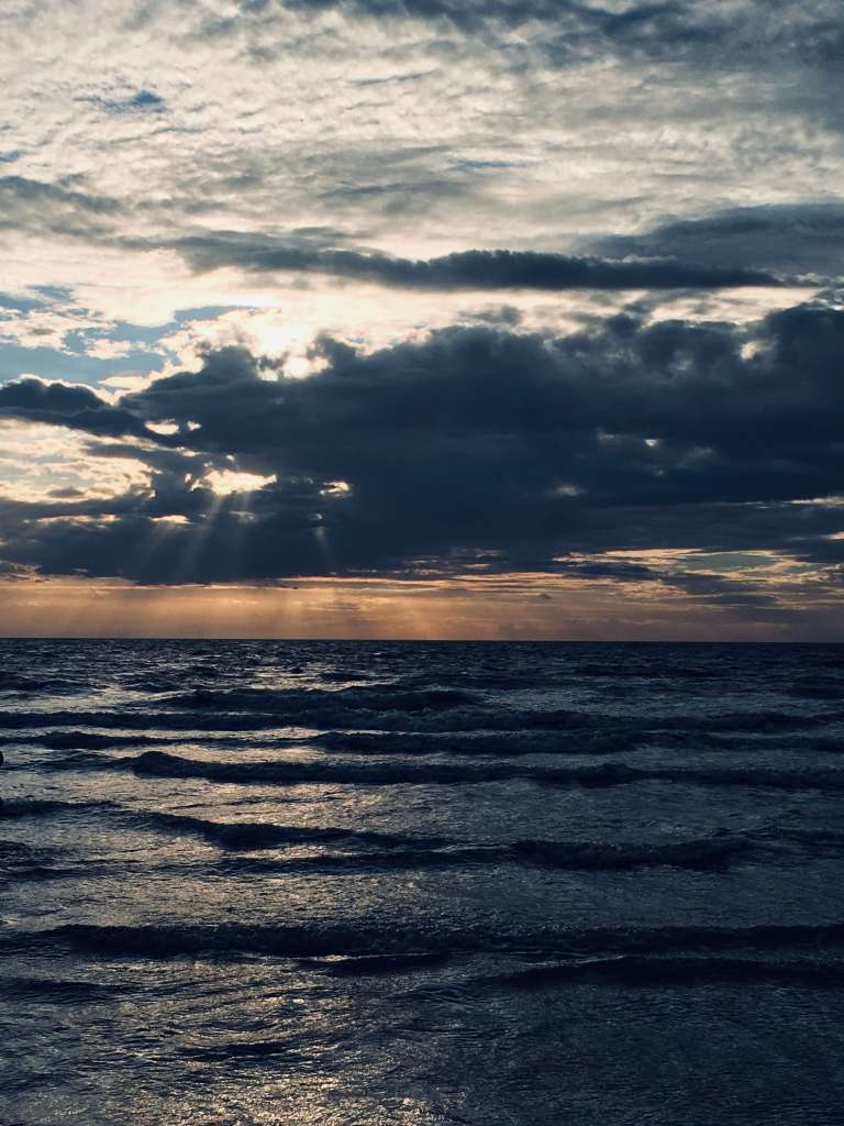 On the horizon, the glow of late evening sunlight is visible behind the clouds, shining onto dark and glittering waves as the tide comes in across a beach
