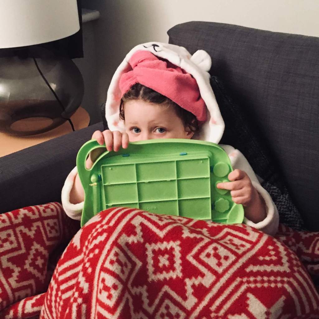 A child sits on a sofa in her dressing gown, with a magnetic drawing board propped on her knees