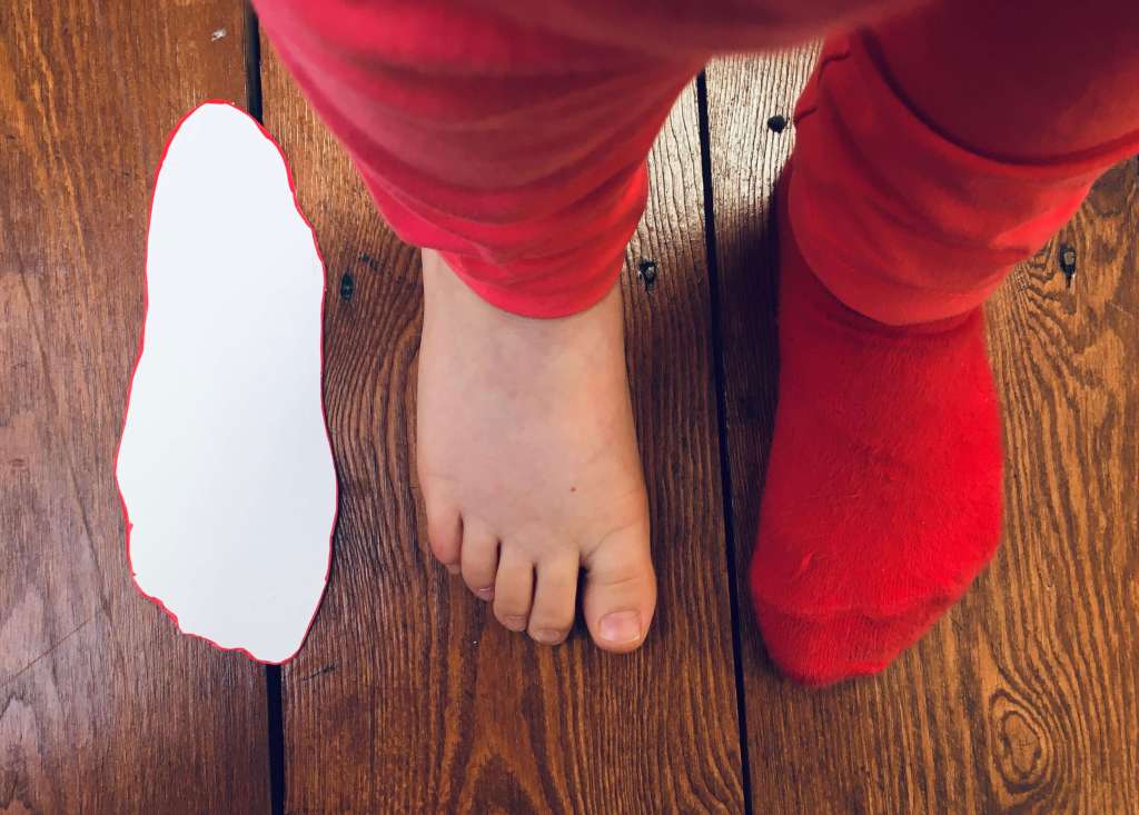 A child's feet are shown next to a cardboard template measure that was completed by drawing around her foot