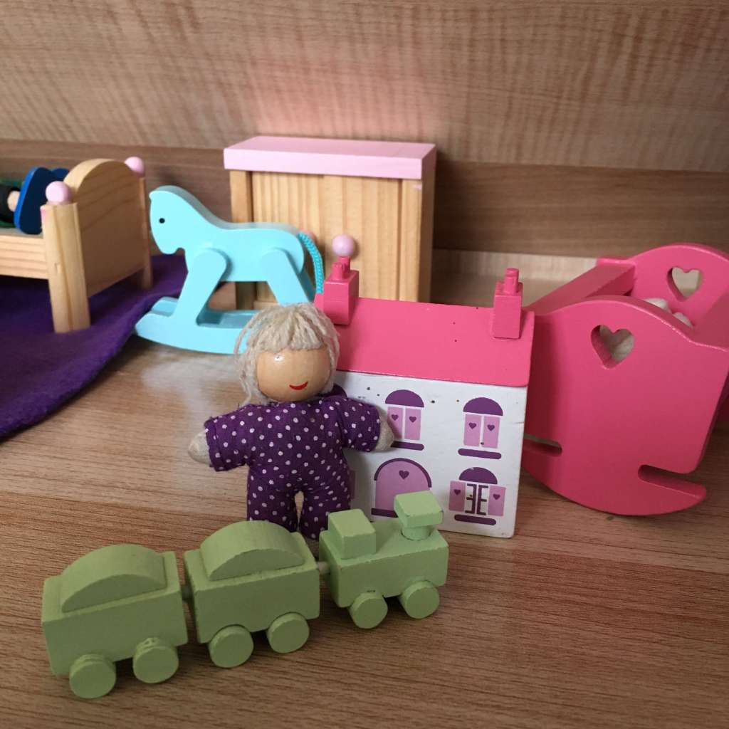 A wooden baby doll next to a tiny replica wooden doll's house, a tiny wooden train set and her little wooden crib, all within a large doll's house