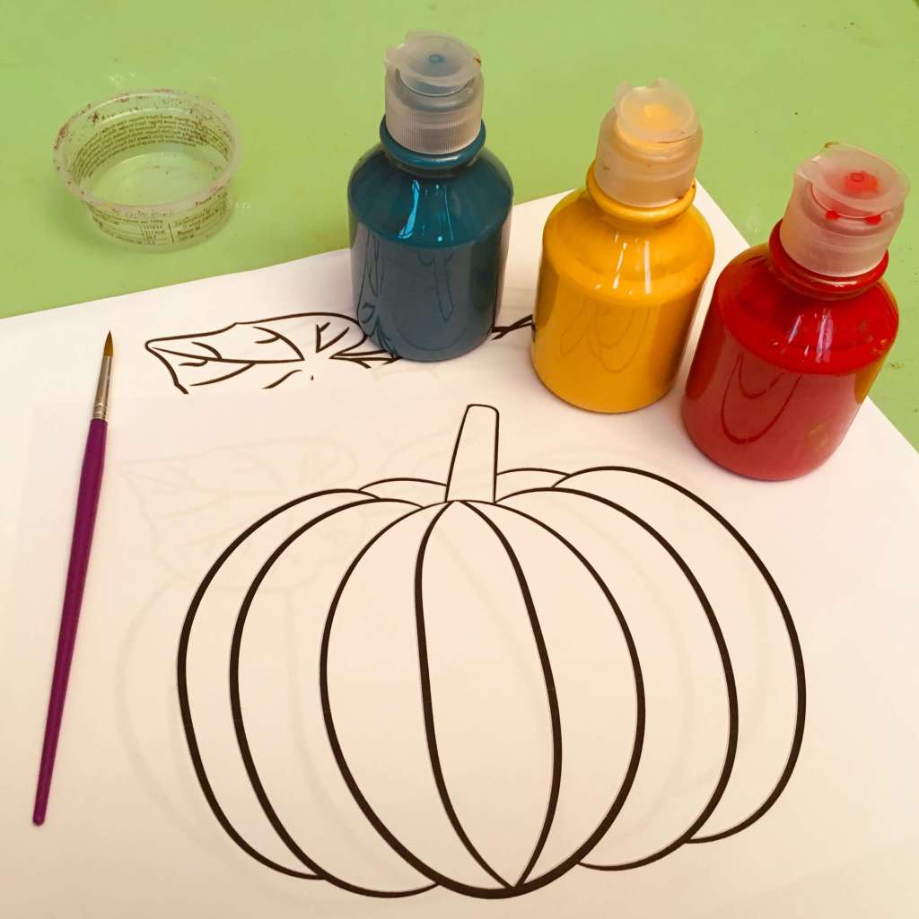 Three pots or primary colour poster paints, a paint brush and some printed paper images of pumpkins, ready to be painted