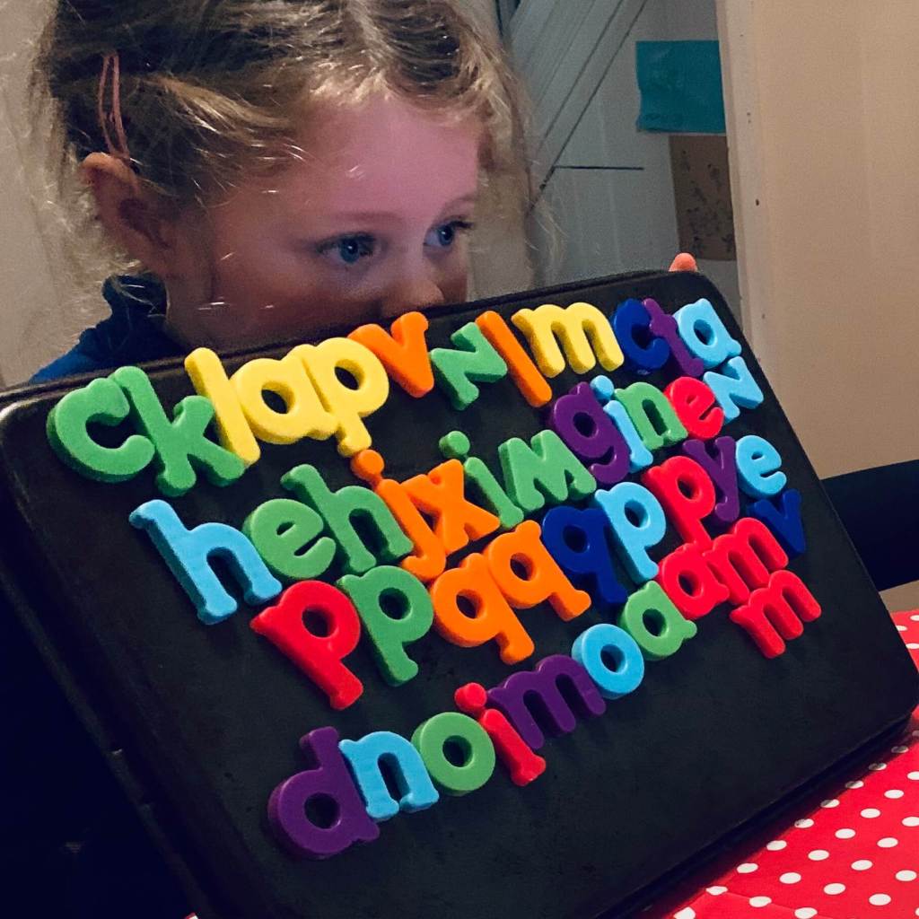 A girl holds up a metal baking tray covered by magnetic letters