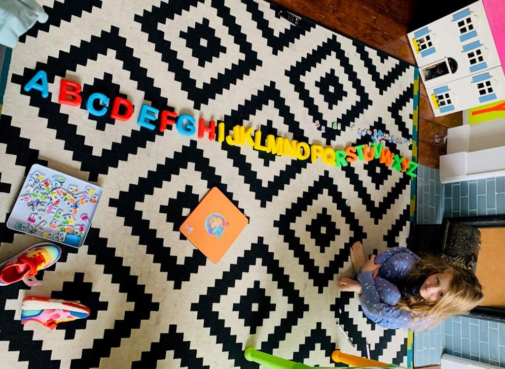 A child sits next to a row of colourful uppercase letters arranged on the floor, forming the Latin alphabet