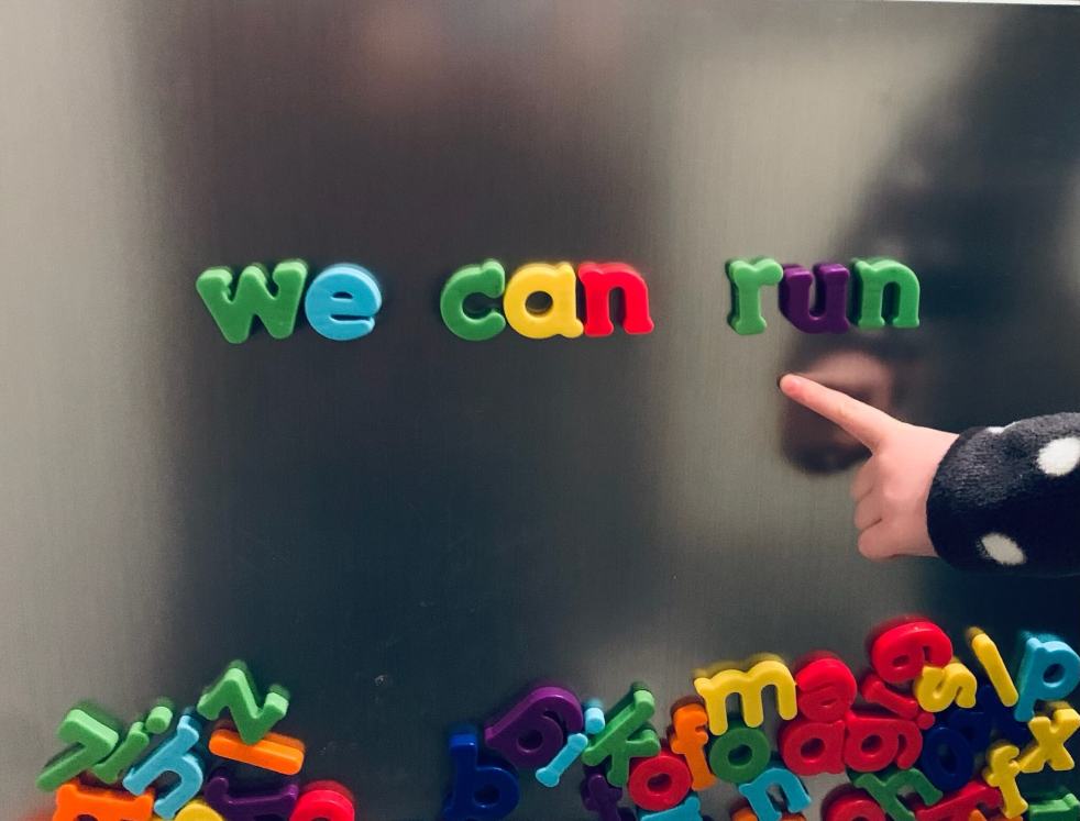 The sentence 'we can run' written in magnetic letters on a fridge door, with a child pointing to the words