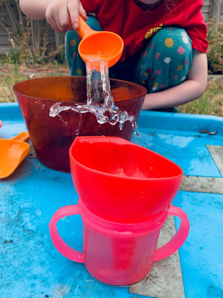 A picture of a young child undergoing a STEM activity, involving transferring water from a large bowl into a smaller cup using a variety of tools: spoons, scoops, a pipette, a funnel and a jug.