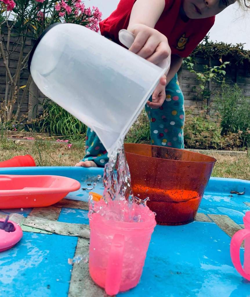 A picture of a young child undergoing a STEM activity, involving transferring water from a large bowl into a smaller cup using a variety of tools: spoons, scoops, a pipette, a funnel and a jug.
