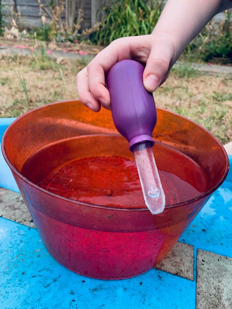 A picture of a young child undergoing a STEM activity, involving transferring water from a large bowl into a smaller cup using a variety of tools: spoons, scoops, a pipette, a funnel and a jug.