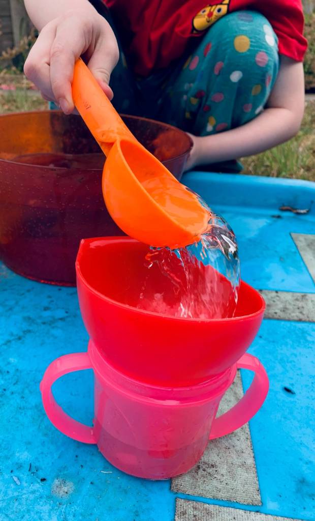 A picture of a young child undergoing a STEM activity, involving transferring water from a large bowl into a smaller cup using a variety of tools: spoons, scoops, a pipette, a funnel and a jug.