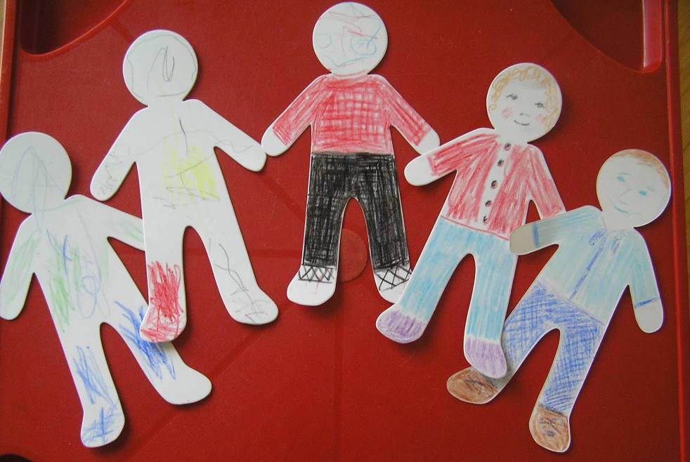 A group of five paper dolls (or cut outs) some coloured by a child and some by adults, shown on a red tabletop