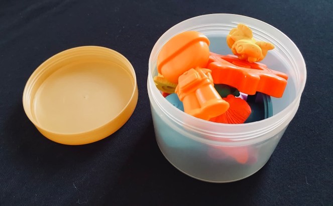 A small translucent pot with the lid off filled with multicoloured toys, on a black background